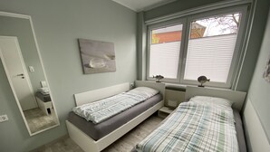 A bedroom with two single beds, a bedside table with a lamp, a window with blinds, and a wall-mounted painting.