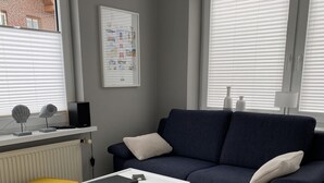 A modern living room with a dark blue sofa, a white coffee table, and a yellow ottoman. A framed picture hangs on the wall, and there are two windows with blinds.