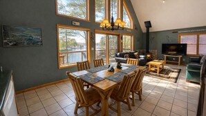 Un espace repas avec une table et des chaises en bois, une cheminée et de grandes fenêtres donnant sur des arbres et un lac.