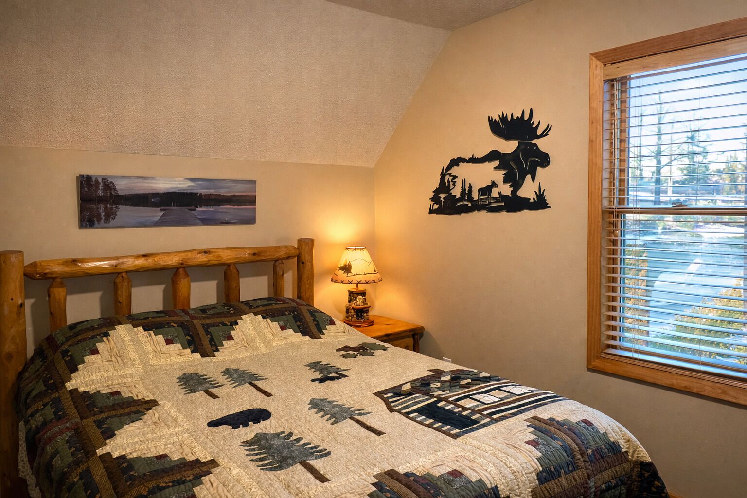 A bedroom with a wooden headboard, a quilt with prehistoric animal patterns, a lamp, a wall decal of a moose, a window with blinds, and a painting of a landscape.