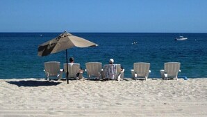 A beach with four white chairs under a beige umbrella, facing the ocean. - Casa Cardonal - El Cardonal, BCS Mexico (El Cardonal)
