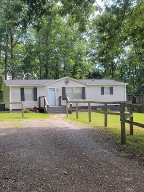 A two-story mobile home with a front porch and a small deck, surrounded by a wooden fence and trees. - Indian Creek Cabin - A welcome retreat. (Tryon)