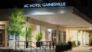 Das moderne Äußere des AC Hotels Gainesville mit einem markanten Eingang, großen Kübelpflanzen und einer gut beleuchteten Fassade.