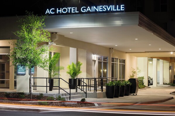 Het exterieur van het AC Hotel Gainesville, met een modern ontwerp, met een opvallende ingang, grote potplanten en een goed verlichte gevel.