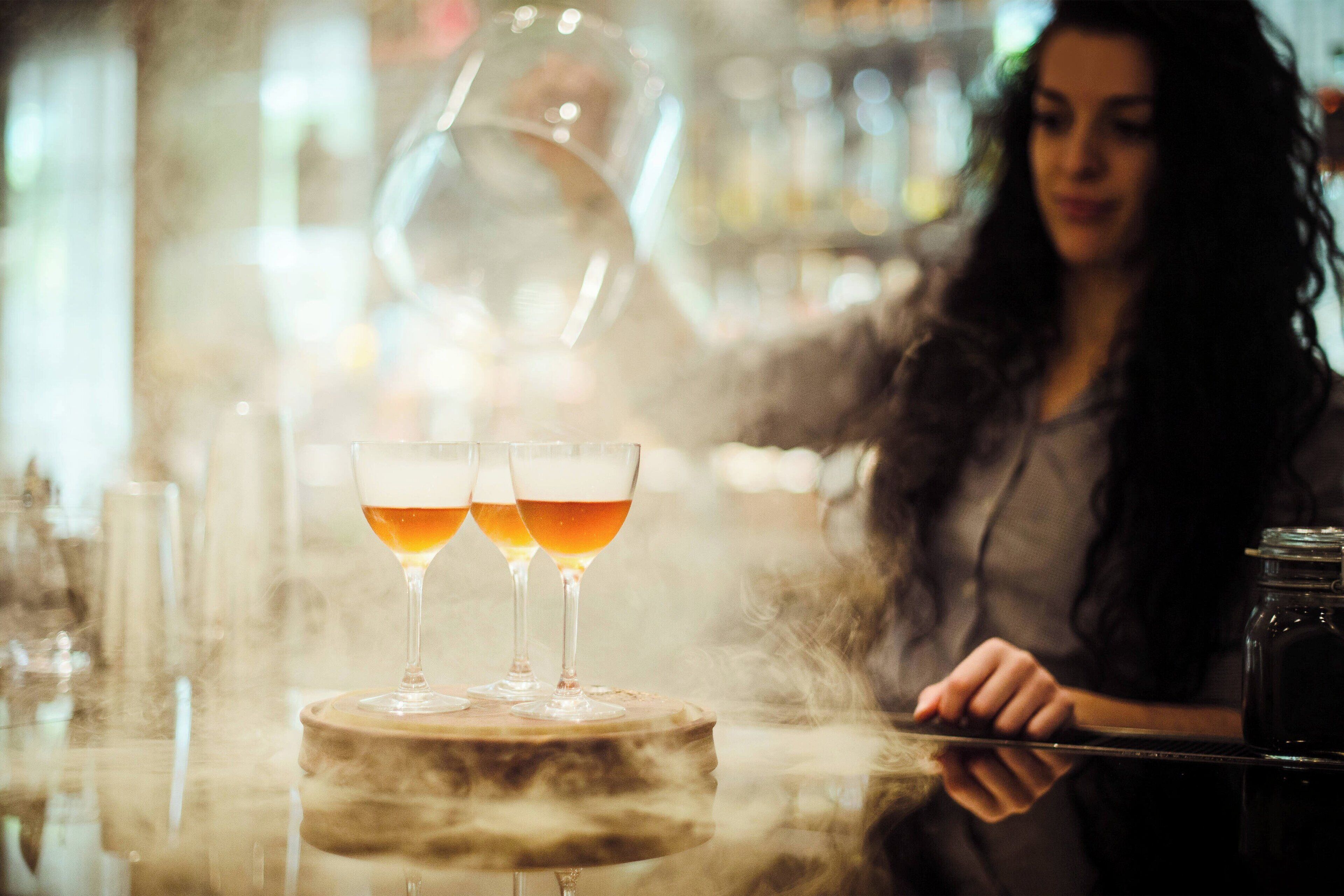 A bar setting with two glasses of amber-colored beverage on a round table, a person in a dark outfit behind the bar, and a jar on the counter.