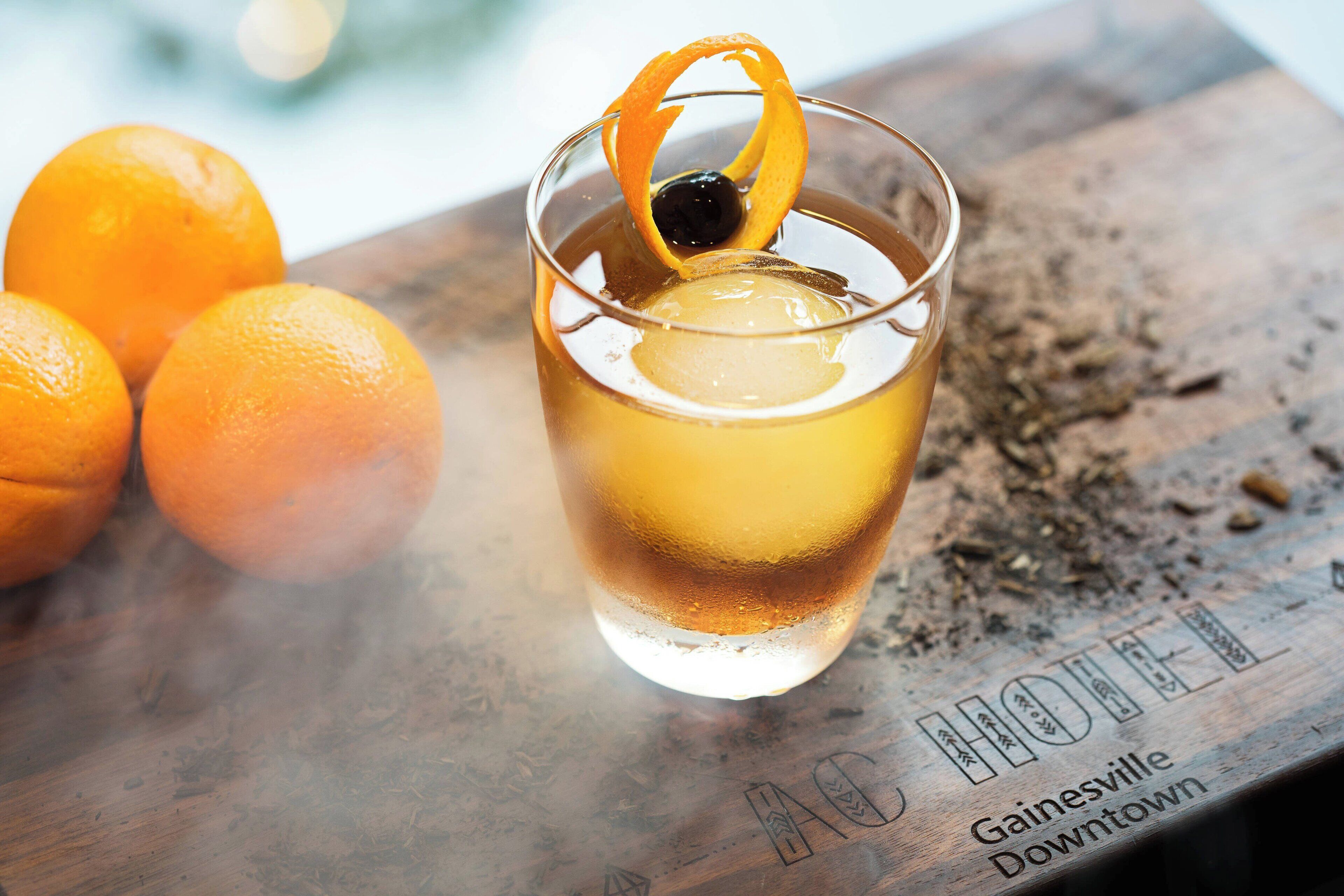 Un verre contenant une boisson de couleur ambrée, garni d’un zeste d’orange et d’une olive noire, posé sur une surface en bois, avec l’inscription « Gainesville Downtown » visible.