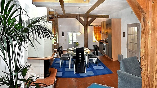 A spacious living area with a dining table, chairs, a sofa, and a blue rug. There is a kitchen area with cabinets and a sink. The room has wooden beams and a balcony with a railing. - Spacious vacation residence for 6 pers. with pool and garden. Sauna - shared use (Stadland-Reitland)