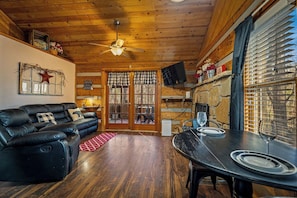 L’intérieur chaleureux d’un chalet avec un plafond en bois, un canapé en cuir noir, une table à manger avec des chaises et un téléviseur fixé au mur.