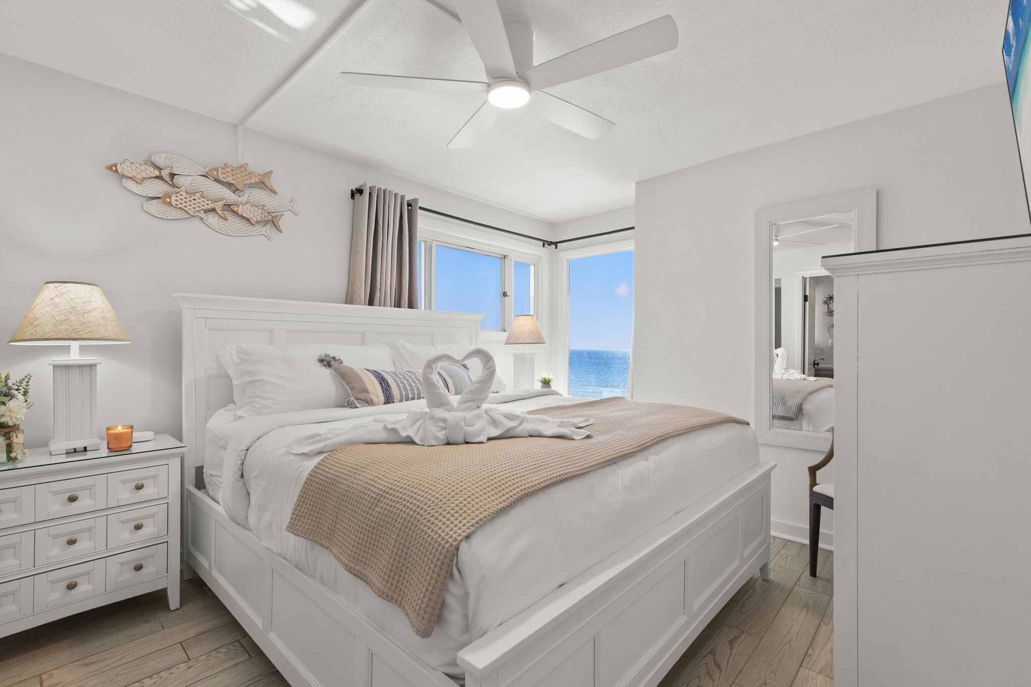 A bedroom with a bed, nightstands, a ceiling fan, a window with curtains, and a view of the ocean.