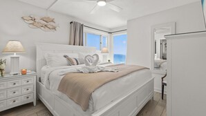A bedroom with a bed, nightstands, a ceiling fan, a window with curtains, and a view of the ocean. - Gulf-Front Comfort, Personally Prepared for You (Panama City Beach)