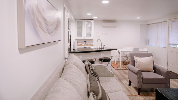 A modern living room with a sofa, armchair, and a small coffee table. There is a kitchen area with a sink and cabinets in the background.