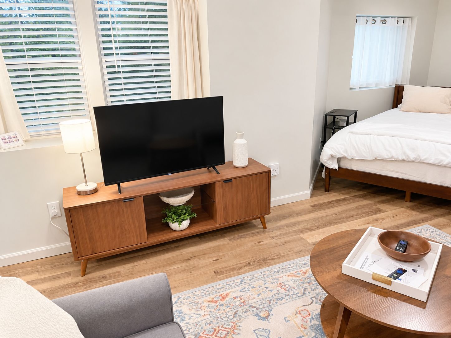 A modern hotel room with a flat-screen TV on a wooden console, a bed with white bedding, a bedside table with a lamp, and a small potted plant.