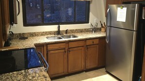 A kitchen with wooden cabinets, a stainless steel refrigerator, a granite countertop, a double sink, and a window with a view of trees.