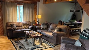 A cozy living room with a wooden ceiling, leather sofas, a coffee table, and a dining area with a plant.