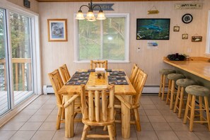 Een houten eettafel met stoelen, een bar met krukken, een raam met zonwering en een wandklok.