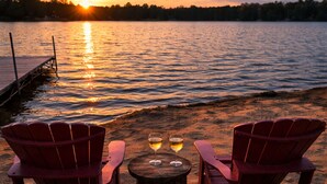 Two red Adirondack chairs on a sandy beach with a wooden table and two wine glasses, overlooking a calm lake at sunset. - Sunset Beach Chalet with Hot Tub & Fire Pit Near Hiking, Skiing & Snowmobiling (Gaylord)
