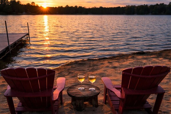 Two red Adirondack chairs on a sandy beach with a wooden table and two wine glasses, overlooking a calm lake at sunset.