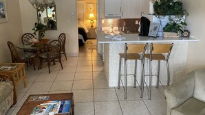 A spacious living area with a dining table, chairs, a coffee table, and a sofa. A kitchen with a white island and bar stools is visible in the background. - BEST DEALS NOW !AMAZING BEACH, PRIVACY, UNWIND. SLEEP 4, FREE CHAIRS, BBQ, PKG  (Madeira Beach)