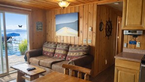 Una cabaña con interior de madera, un sofá con cojines estampados, una mesa, un cuadro de un barco en el agua, un letrero que dice 'Welcome to Lake' y vista de un cuerpo de agua a través de un vidrio.