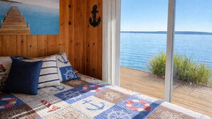 Un dormitorio con una cama grande, ropa de cama con temática náutica, un cuadro de un muelle y vistas a un lago a través de una puerta corrediza de vidrio.