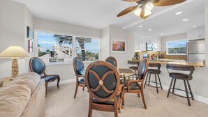 A modern living room with a dining area, kitchen, and large windows with blinds.