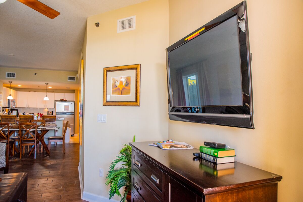 A living room with a flat-screen TV mounted on the wall, a wooden desk with books, and a framed picture on the wall.