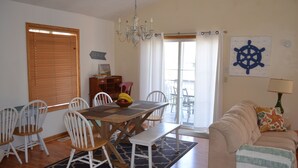Un coin repas avec une table en bois, des chaises blanches et un banc. On y trouve un lustre, un buffet en bois et une fenêtre avec des stores.