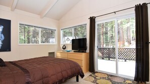 A bedroom with a bed, a TV on a wooden dresser, and large windows with a view of trees.
