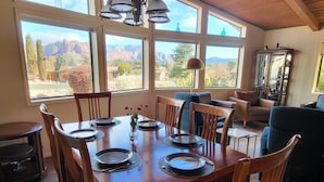 A dining area with a wooden table set for six, large windows with a mountain view, and a dark-colored sofa in the background. - BEST VIEWS! Sleeps 8 Balcony, Firepit, Lawn, Patio, Wildlife (Sedona)
