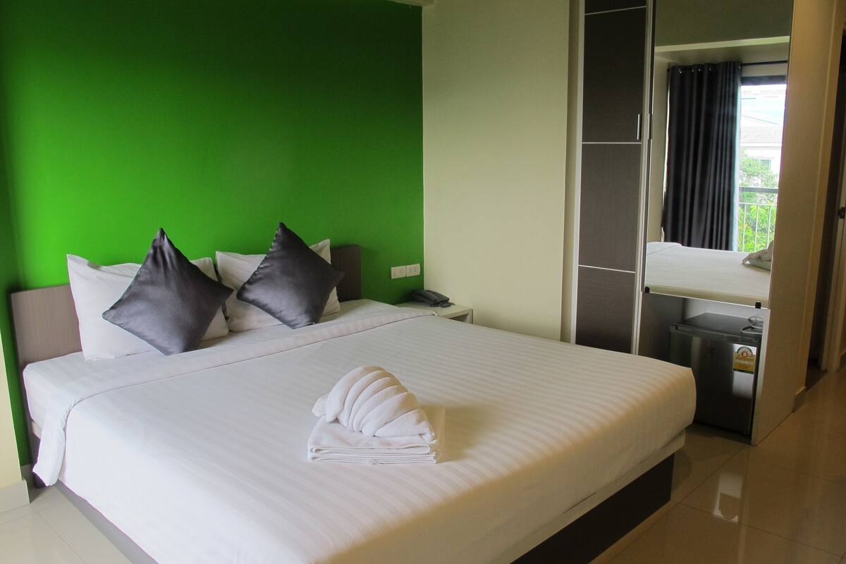 A hotel room with a bed, pillows, a nightstand, a phone, a mirror, and a small trash bin.