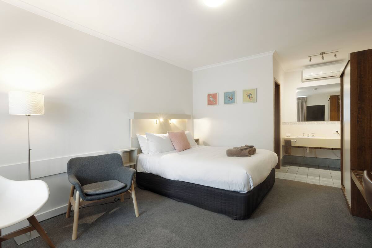 Habitación de hotel con una cama, una silla, un escritorio, una lámpara, un espejo y obras de arte en la pared.