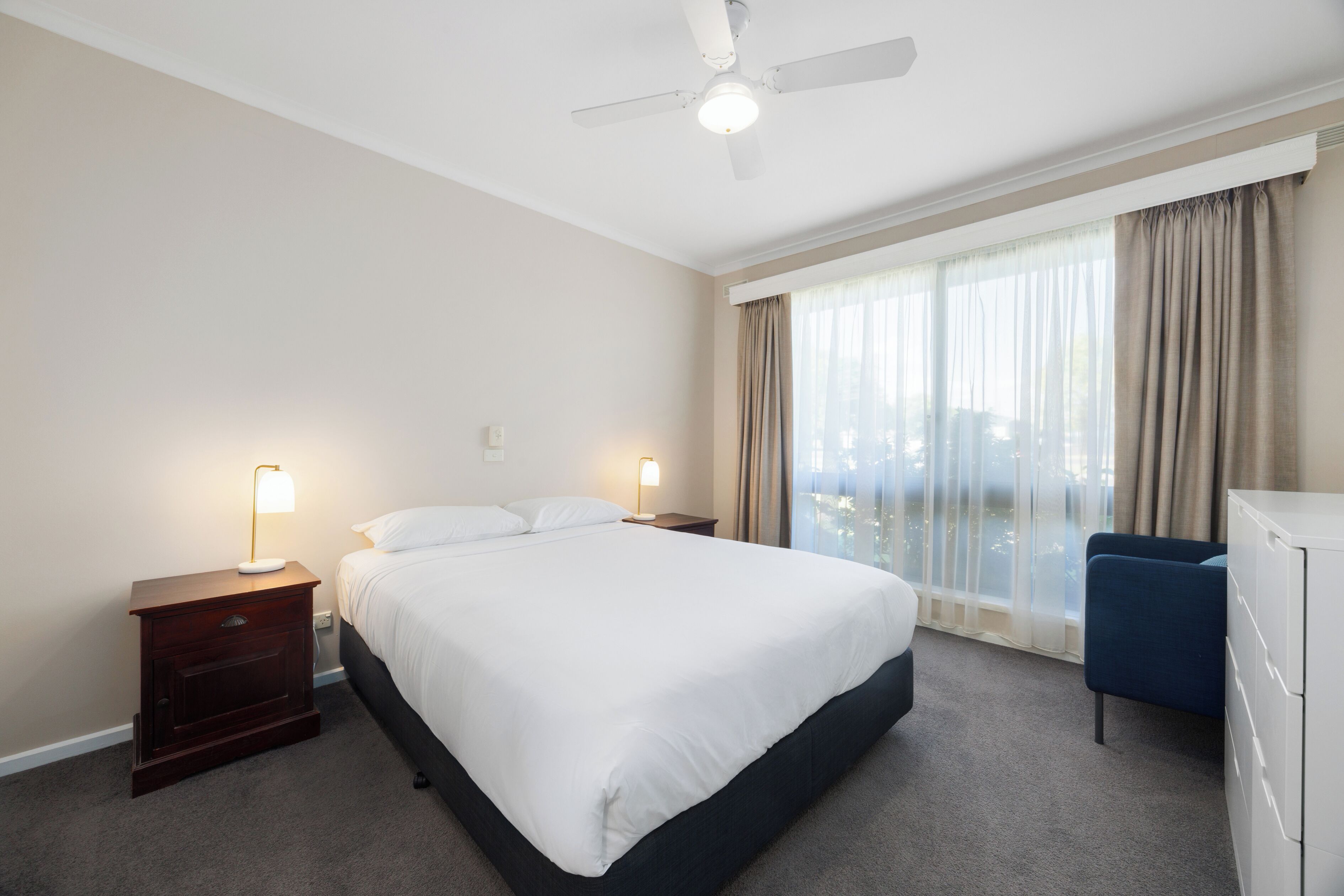 Habitación de hotel con una cama grande, dos lámparas de noche, ventilador de techo, vistas al exterior a través de una ventana y un escritorio con silla.