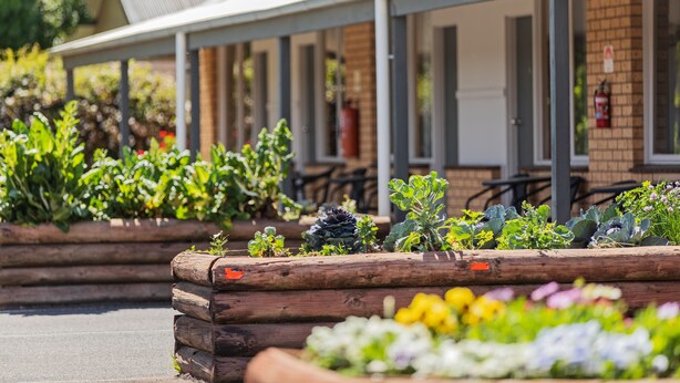 Una fila de macetas de madera con diversas plantas y flores, un edificio de ladrillo con techo metálico, y mobiliario exterior.