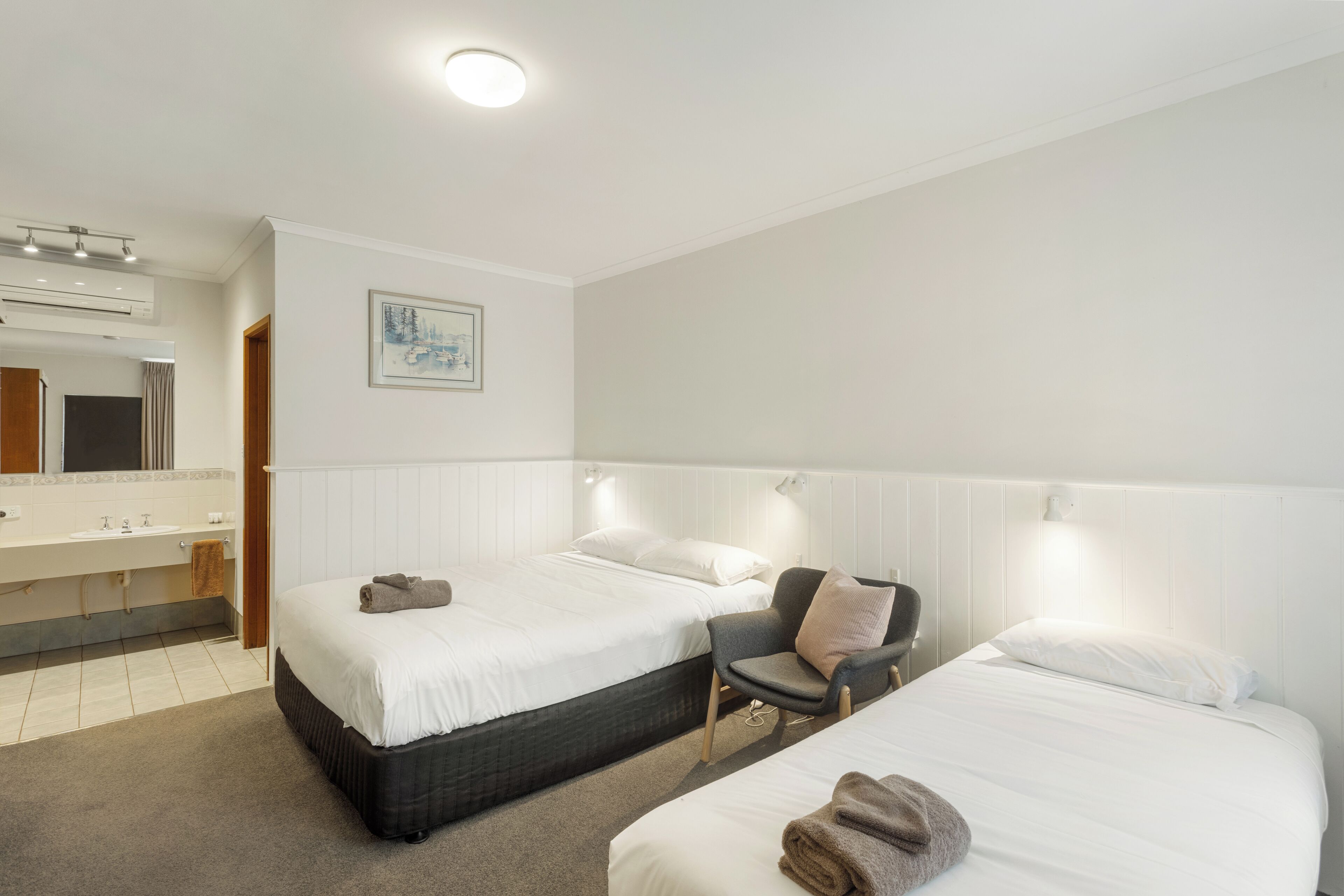 A hotel room with two beds, a bathroom with a sink and mirror, and a painting on the wall.
