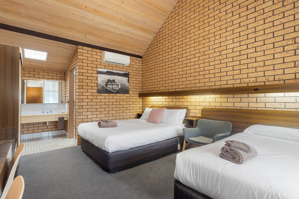 Habitación de hotel con pared de ladrillo, techo de madera, cama con ropa blanca, mesita de noche y una silla.