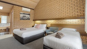 A hotel room with two beds, a wooden ceiling, a brick wall, a painting, a desk, and a chair.