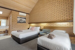 A hotel room with two beds, a wooden ceiling, a brick wall, a painting, a desk, and a chair.