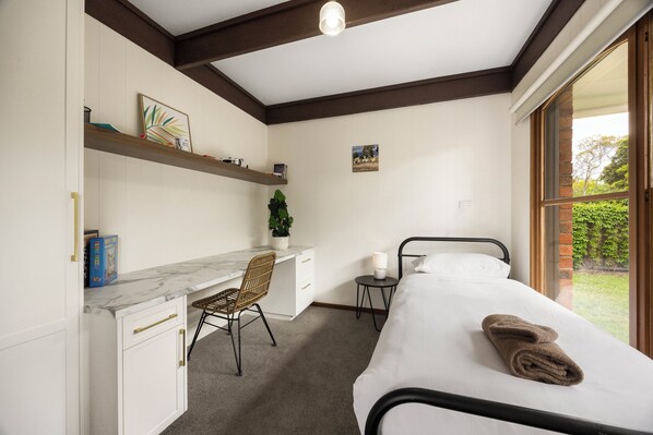 A bedroom with a bed, a desk, a chair, a small table with a lamp, and a window with a view of greenery.
