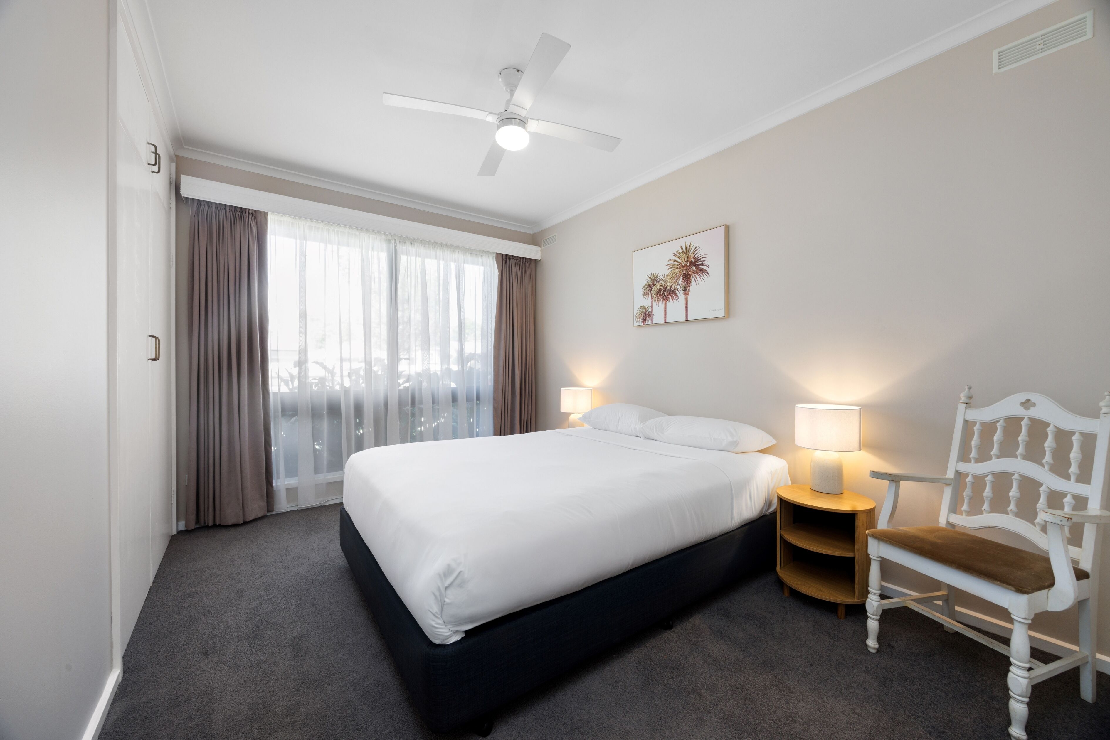 Ein Hotelzimmer mit einem großen Bett, einem weißen Stuhl, einem Deckenventilator, einem Nachttisch mit Lampe und einem Fenster mit Vorhängen.