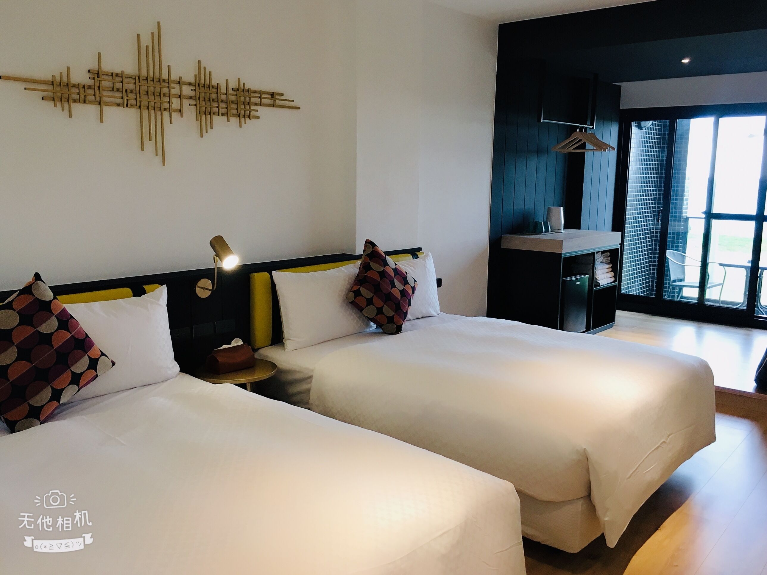 Una habitación de hotel con dos camas, una mesita de noche con lámpara, una obra de arte en la pared y vistas al exterior a través de un ventanal.