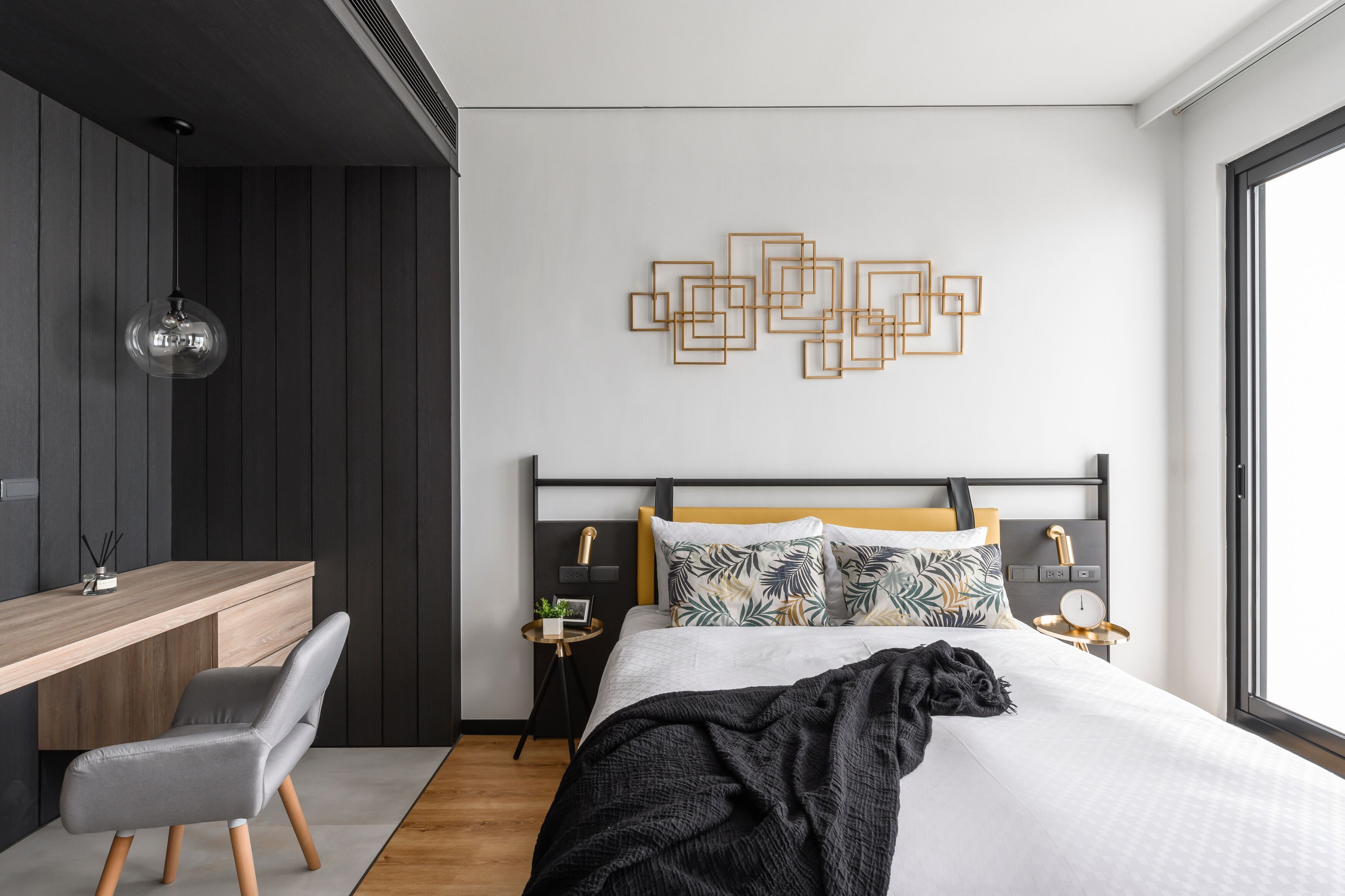 Un dormitorio moderno con un escritorio de madera, una silla gris, una cama con cabecero blanco y una obra de arte geométrico en la pared.