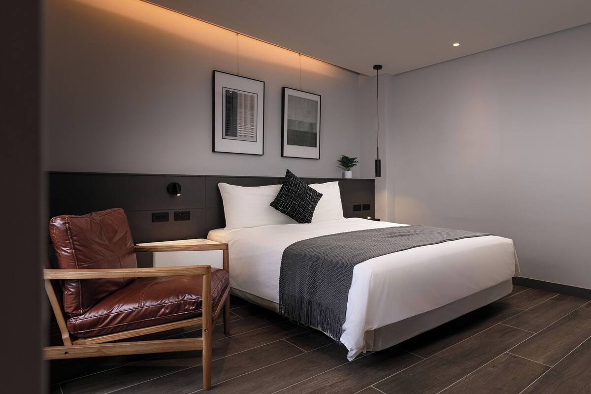 Un dormitorio moderno con una cama, una silla de madera, una mesita de noche y dos cuadros en la pared.
