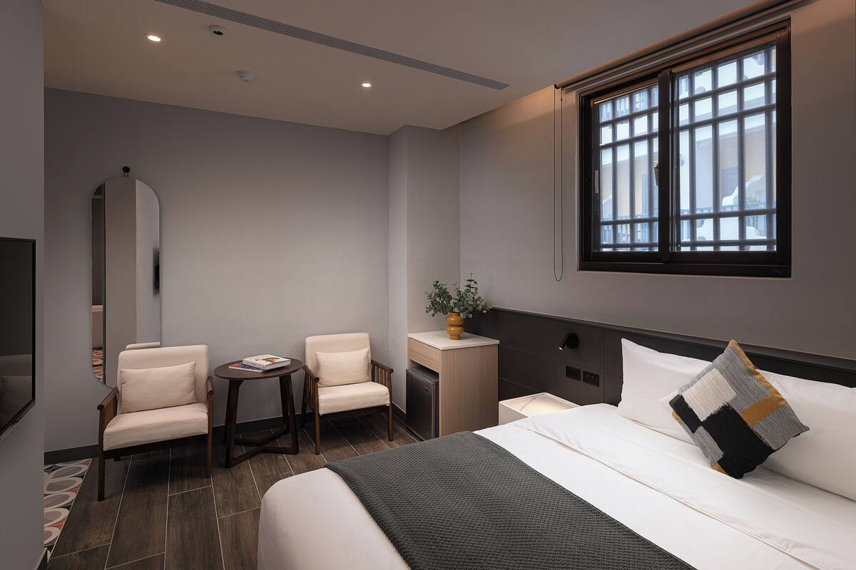 Habitación de hotel con cama, dos sillas, una mesita, un espejo y una ventana.
