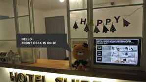 Ein Hotelrezeptionstresen mit einem weichen Bären als Maskottchen, einem 'Happy'-Banner und einem digitalen Display mit Zimmerinformationen.