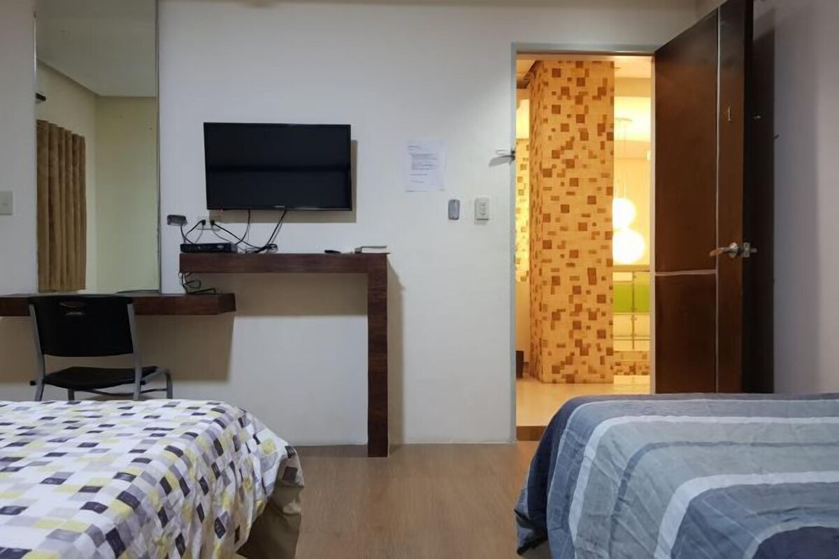 A hotel room with a bed, a desk with a computer, a TV mounted on the wall, and a door leading to another room.