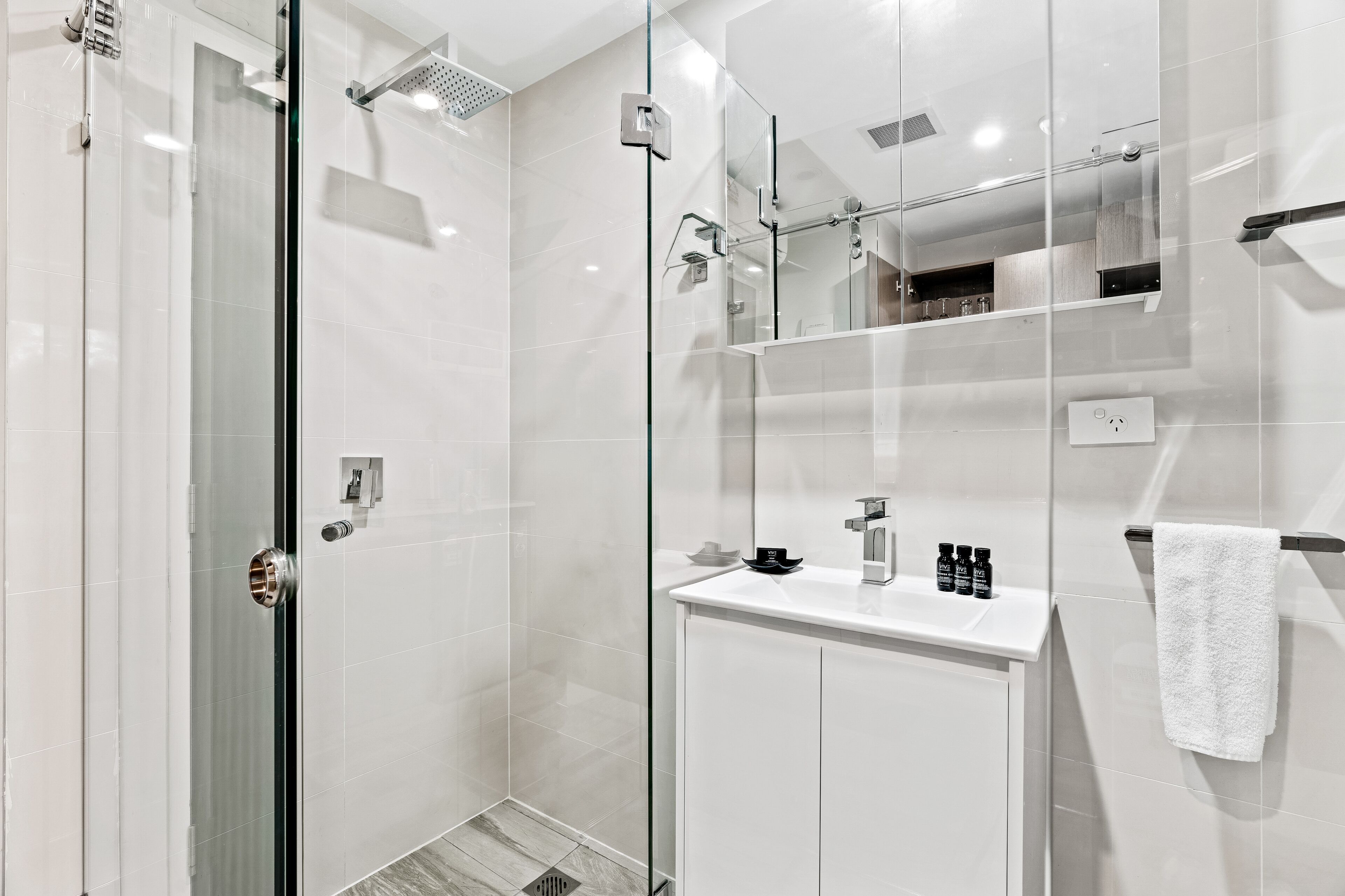 Un baño moderno con ducha envuelta en vidrio, tocador blanco con espejo y una toalla en un perchero.