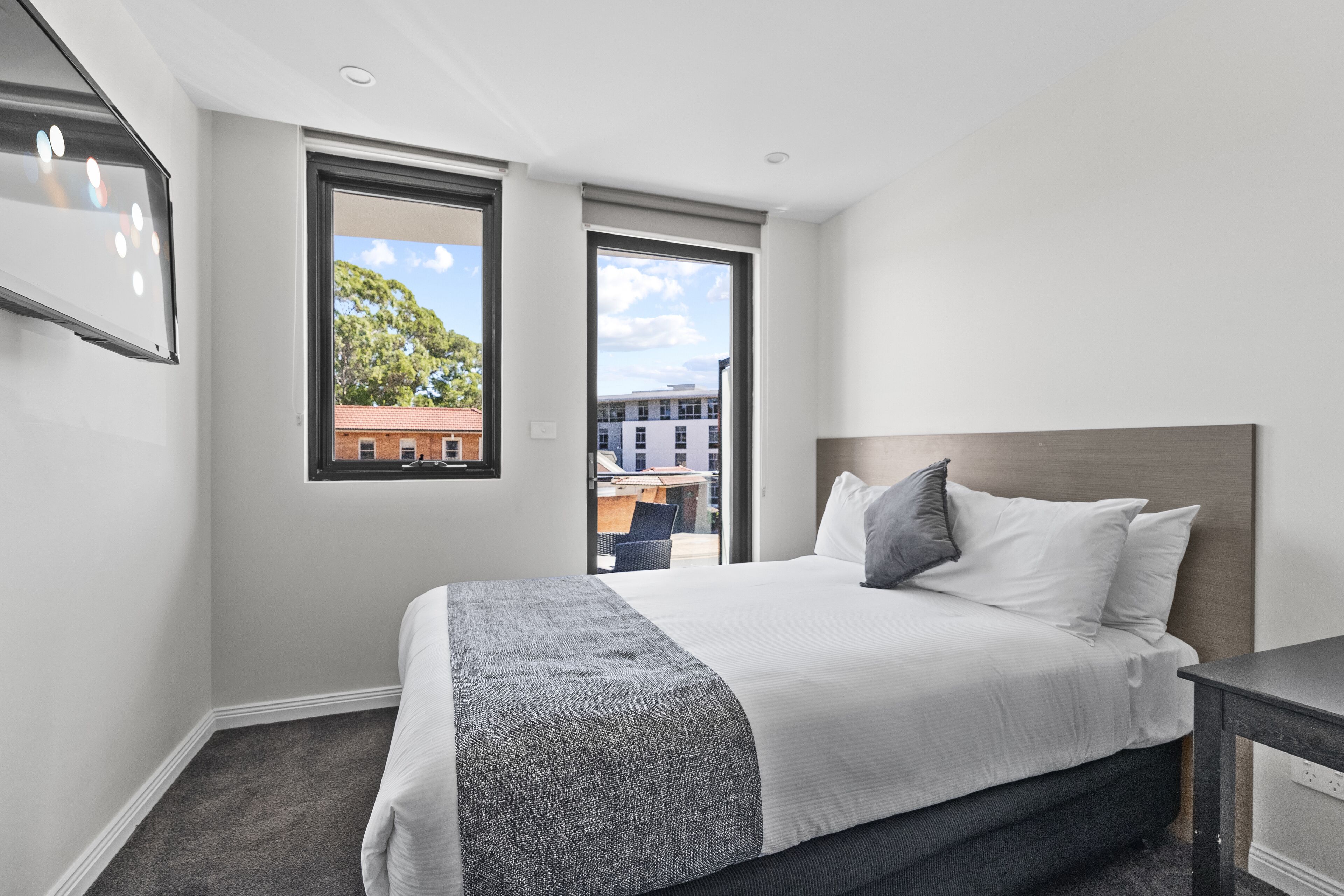 Quarto de hotel moderno com uma cama grande, uma escrivaninha e vista para a cidade pelas janelas.
