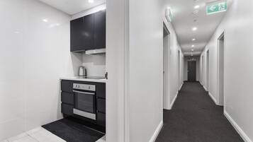 A modern kitchen with white tiles, black cabinets, and a black rug. A hallway with a carpeted floor and white walls leads to a door at the end.
