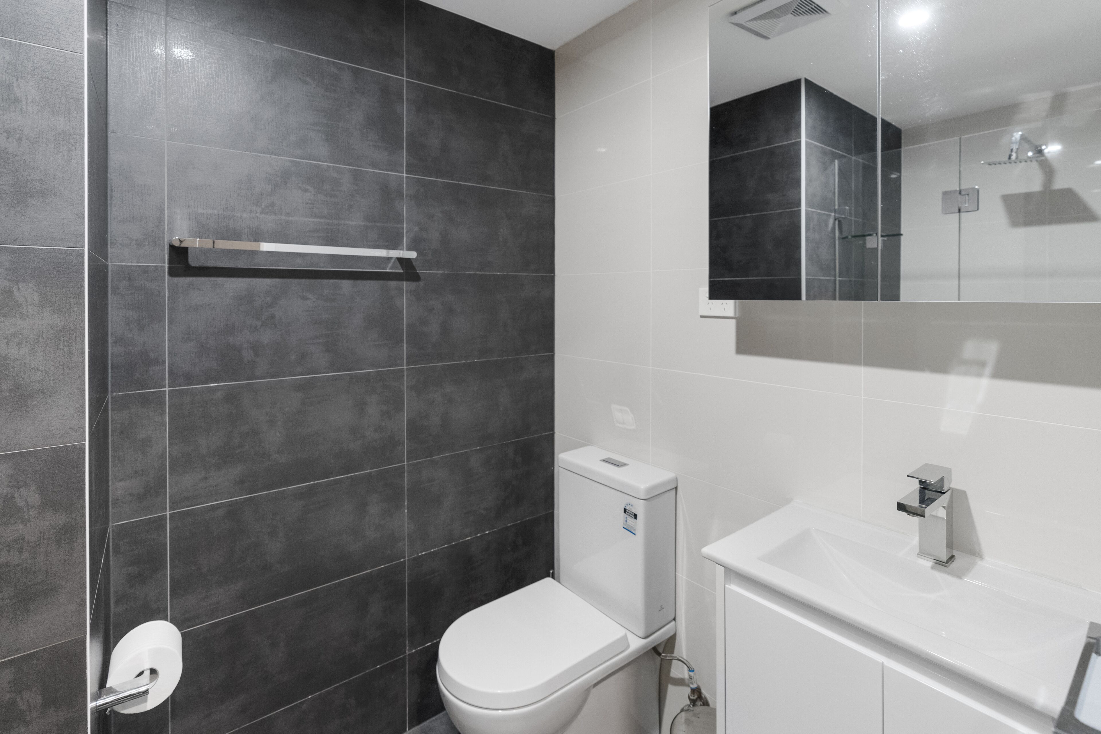 A modern bathroom with dark tiled walls, a white toilet, and a white sink with a mirror above it.