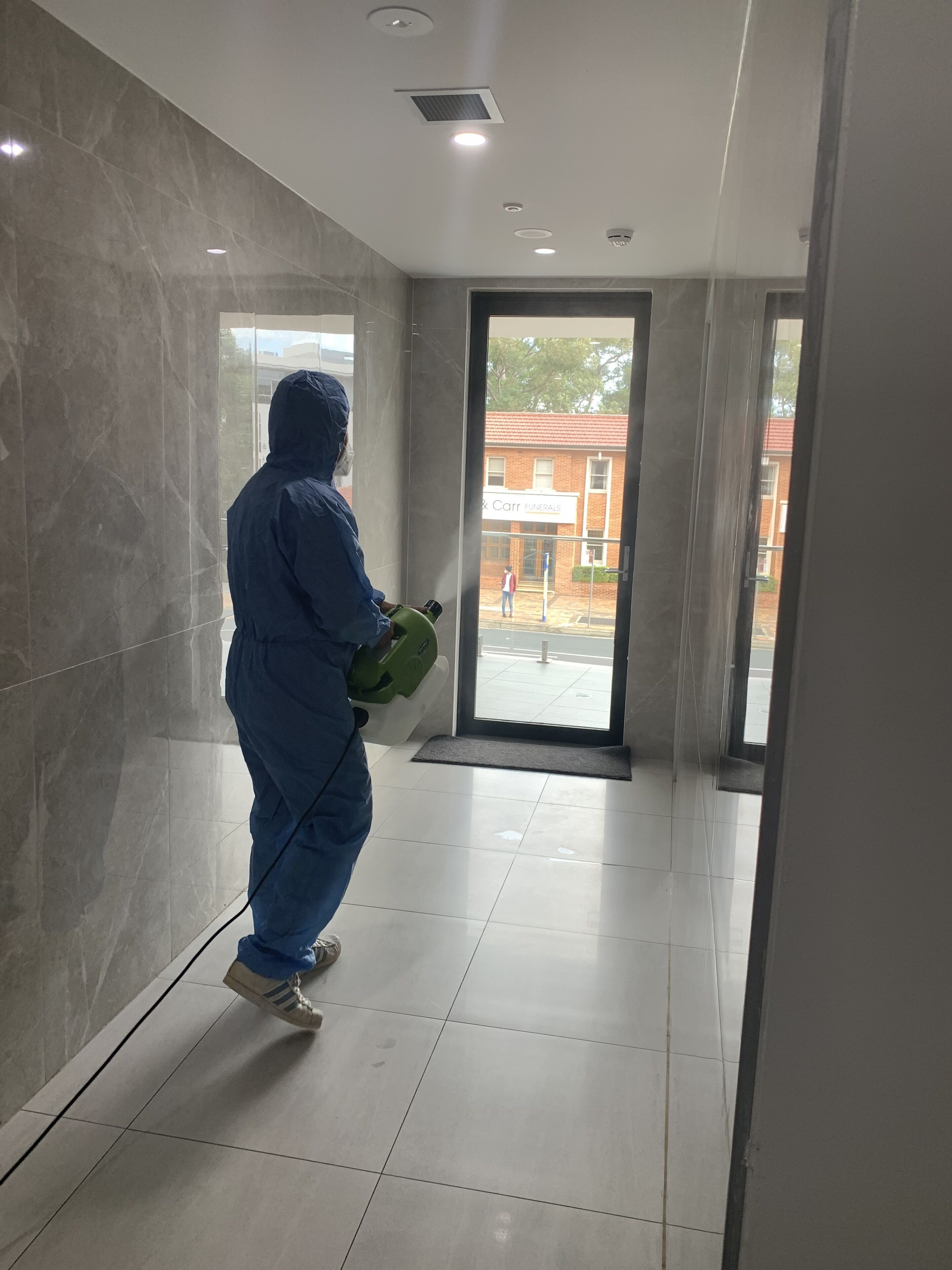 Eine Person in einem blauen Schutzanzug sprüht einen Raum mit einem grünen Gerät ein. Der Raum hat einen gefliesten Boden und eine Tür, die nach draußen führt.